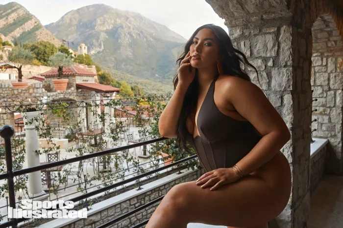 Yumi Nu leans on a railing overlooking the city, wearing a olive one-piece swimsuit.